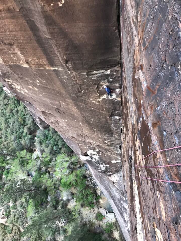 Kippy Hoene standing on a tiny ledge high on the Dark Shadows climbing route, surrounded by sheer rock and a vast drop below.